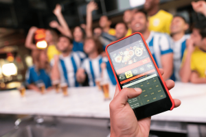 New Jersey lawmaker seeks to ban fast-paced micro betting wagers. Person holding a smartphone with a live sports betting app open, showing football betting options, while a group of excited fans in jerseys cheer in the background at a bar.