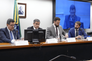 Brazil Congress told to focus on $6.4B gambling revenue losses to the black market. A panel of four men in suits seated at a long desk during a formal congressional session in Brazil. A Brazilian flag and a framed portrait hang on the wall behind them. Nameplates and microphones are positioned in front of each speaker, along with glasses of water. A large screen in the background displays a live feed of the meeting.