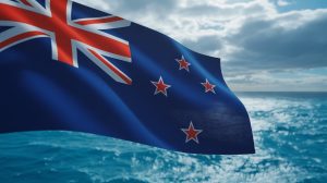 A photograph capturing a close-up of the New Zealand flag rippling dramatically in a strong wind. The flag's dark blue field and four red stars are sharply defined against a cloudy sky. The silver fern emblem on the flag is subtly visible. The backdrop is a vibrant blue ocean under diffused sunlight, adding depth and movement to the scene.