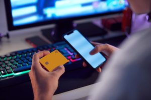 GambleAware shares concern as gambling harm figures double. Person holding a yellow credit card and a smartphone in front of a computer setup with a colorful backlit keyboard and monitor, suggesting online payment or digital transaction activity.