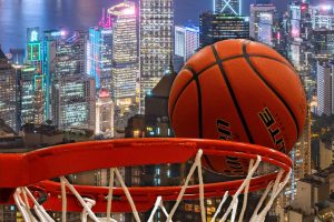 Hong Kong basketball gambling gains support from 77 councillors. A basketball going through a hoop with the illuminated skyscrapers of Hong Kong in the background, symbolizing the intersection of basketball and gambling culture in the city.