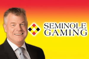 Seminole Gaming's COO speaks out against rise in illegal gambling in Florida. David Hoenemeyer in a black suit, white shirt and grey tie, smiling. Seminole Gaming logo beside him on top of orange and yellow background.