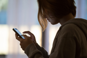 A photograph taken from behind showing a young person holding a smartphone in their hands, their silhouette defined against soft ambient lighting. EGBA launches influencer marketing pledge, with minors to be protected