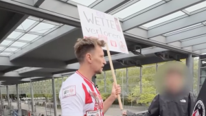Man stood wearing a German football top and holding a sign, with a person stood next to him with their face blurred out.