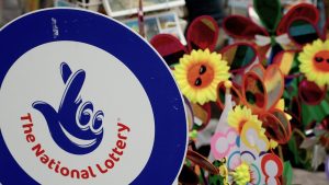 A sign featuring "The National Lottery" logo with a blue background and a hand symbol with crossed fingers. In the background, there are colorful pinwheels and sunflowers with smiling faces, likely part of a festive street stall or market. UK's National Lottery to temporarily shut down for major tech updates. UK's National Lottery returns to '90s 'It Could Be You' tagline
