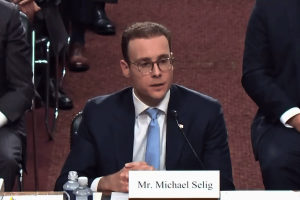 CFTC nominee Michael Selig dodges senators' grilling, keeping sports prediction markets in legal limbo. Michael Selig sits at a Senate hearing table, wearing a dark suit, light blue tie, and glasses, speaking into a microphone with a nameplate reading “Mr. Michael Selig” in front of him.