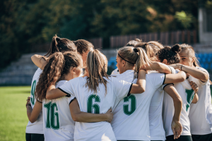 How is the rise of women's soccer impacting sportsbooks. A women’s soccer team stands in a tight huddle on the field, arms around each other, wearing white jerseys with green numbers. They appear focused and unified, preparing together before a match, with a blurred stadium and trees in the background.