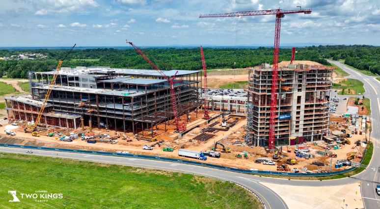 Aerial view of a large construction site with two multi-story buildings under construction, surrounded by cranes, construction vehicles, and materials. A curved road borders the site in the foreground, with green fields and trees extending into the distance under a partly cloudy sky.