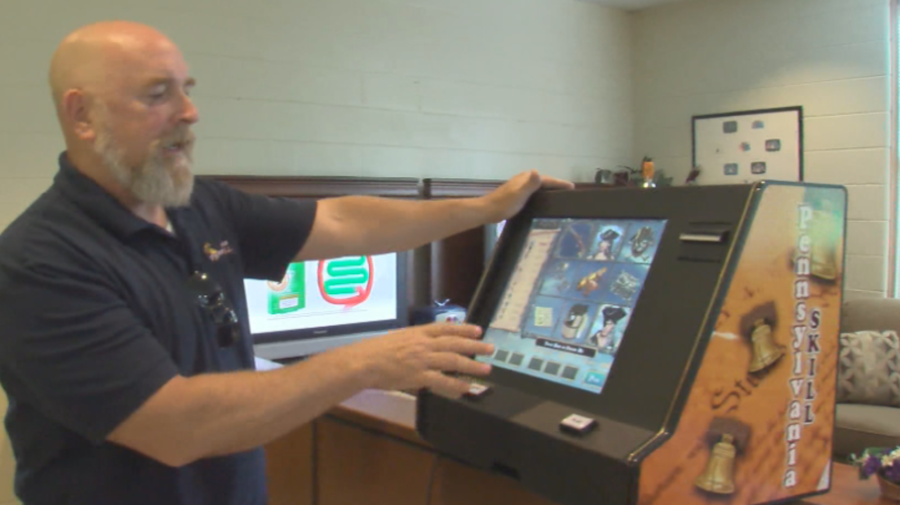 Rick Goodling is a bald, bearded man in a dark polo shirt, demonstrates a video gambling or skill game machine inside an office setting.